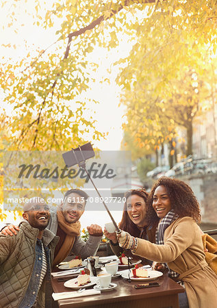 Smiling friends taking selfie with selfie stick at autumn sidewalk cafe