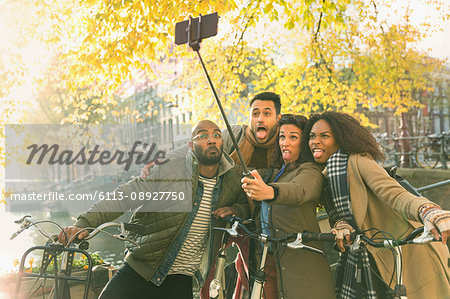 Playful young friends with bicycles making a face taking selfie with selfie stick along autumn canal, Amsterdam