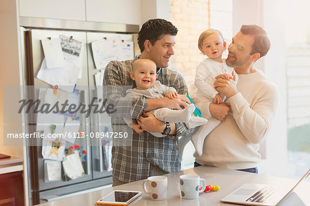 Male gay parents holding baby sons in kitchen