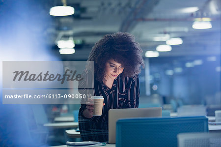 Businesswoman working late at laptop, drinking coffee in dark office