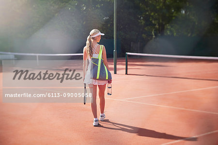 Young female tennis player walking with tennis racket, bag and water bottle on sunny clay tennis court