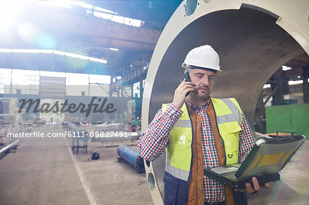 Male engineer with laptop talking on cell phone in steel factory