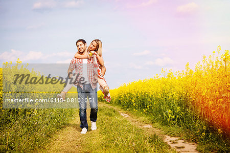 Young man carrying girlfriend piggyback along field path by colza field, Tuscany, Italy