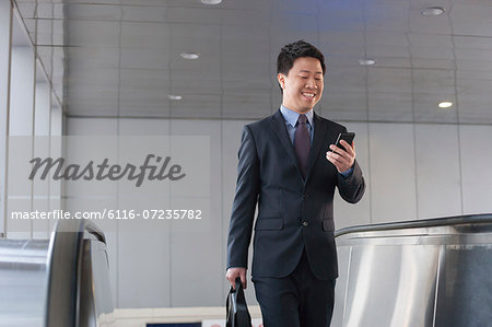 Smiling businessman coming up the escalator and looking down at his phone