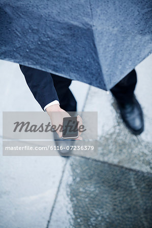 High angle view of businessman holding an umbrella and looking at his phone in the rain