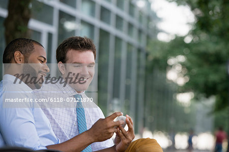 Summer. Two Men Sitting On A Bench, Using A Smart Phone.