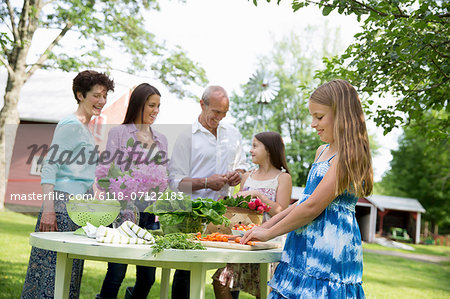 Family Party. Five People Gathered Around A Table Preparing Fresh Salads And Fruit For A Party. Two Girls, One Young Woman And A Mature Couple.
