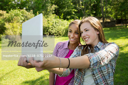 Two Girls Sitting Outdoors On A Bench, Using A Digital Tablet. Holding It Out At Arm's Length.