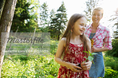 A Young Girl Holding A Seedling Plant, And Walking Through The Gardens Of The Farm With An Adult Woman Following.