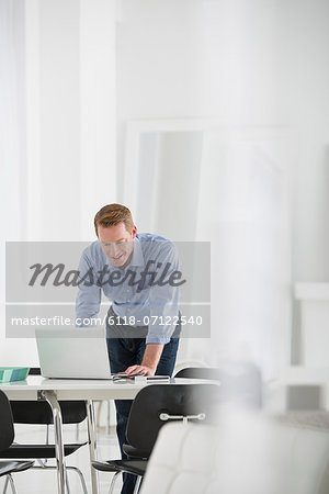 Business. A Man Standing Over A Desk, Leaning Down To Use A Laptop Computer.