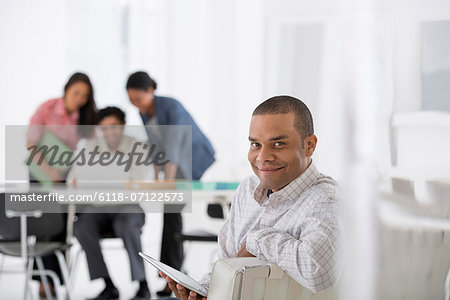 Business. A Man Seated In The Foreground, And Three People Working At A Laptop. Business. A Man Seated In The Foreground, And Three People Working At A Laptop.