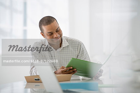 Business. A Man Seated At A Desk Opening A File Of Paperwork. A Laptop Open. Business. A Man Seated At A Desk Opening A File Of Paperwork. A Laptop Open.