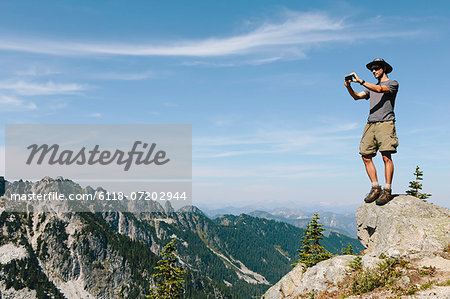 A hiker on a mountain summit, holding a smart phone, at the top of Surprise Mountain, in the Alpine Lakes Wilderness, in Mount Baker-Snoqualmie National Forest.