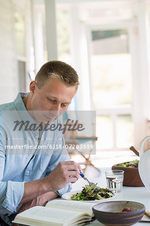 A man seated at a cafe table eating a salad and reading a book.
