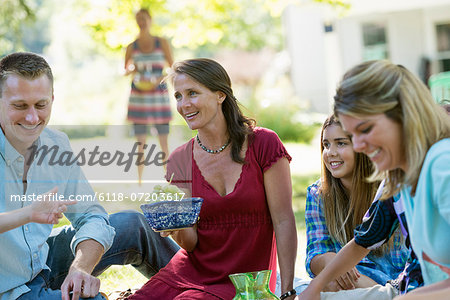 A group of adults and children sitting on the grass under the shade of a tree. A family party.