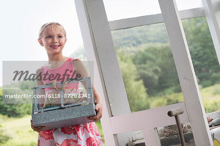 A young girl in a kitchen wearing a pink dress. Carrying a terrarium containing small plants.