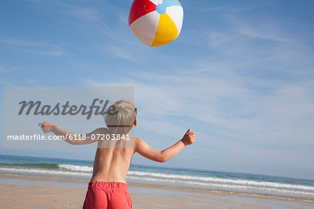 A boy in swimming trunks on the beach, with a large beach ball in the air above him.