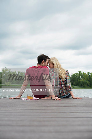 A man and woman seated on a jetty by a lake.