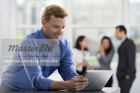 Young professionals at work. A man in an open necked shirt using a digital tablet. A group of men and women in the background. Young professionals at work. A man in an open necked shirt using a digital tablet. A group of men and women in the background.