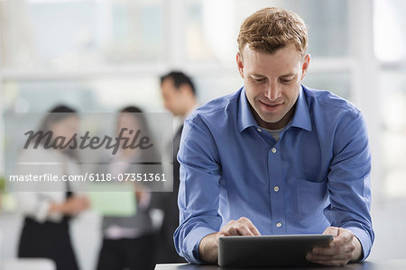 Young professionals at work. A man in an open necked shirt using a digital tablet. A group of men and women in the background. Young professionals at work. A man in an open necked shirt using a digital tablet. A group of men and women in the background.