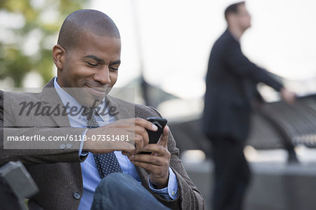 Business people in the city. Keeping in touch on the move. A man seated using his smart phone. A man in the background.