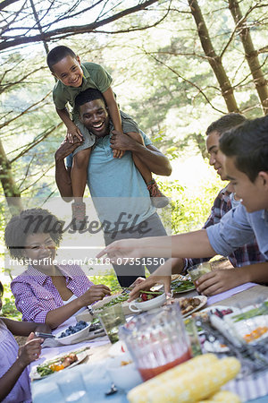 A family picnic meal in the shade of tall trees. A young boy sitting on his father's shoulders.