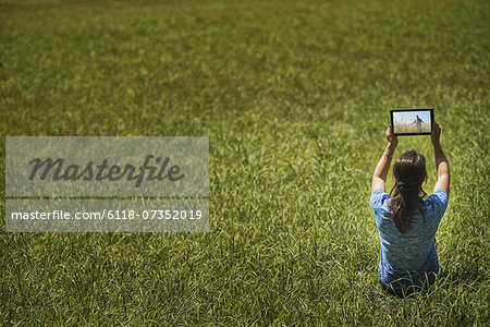 View from above of a woman sitting on grass, lifting up a personal computer notepad, looking at an image.