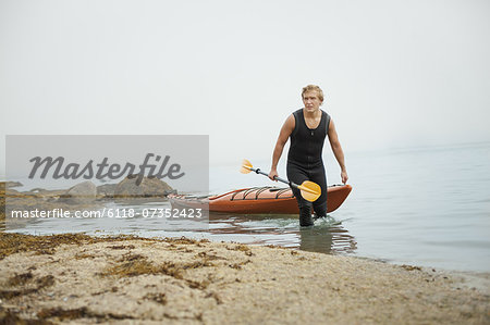 A man in a wetsuit drawing his kayak onto the shore, in misty weather. New York State, USA