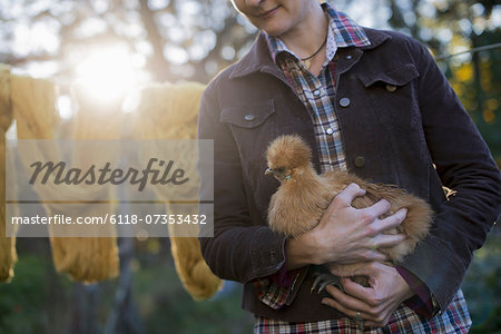 A woman holding a small brown fluffy chicken. A long brown knitted scarf on a washing line. Autumn sunshine filtering through trees.