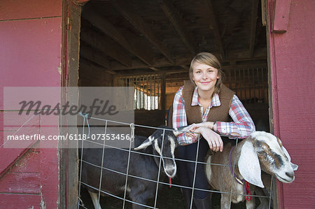 A goat farm. A young girl leaning on the barrier of the goat shed, with two animals peering out.
