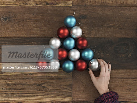 A collection of blue, red and silver ornaments arranged in a triangular shape on a wooden board. A Christmas tree shape. A person's hand placing the final ball.