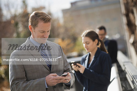 Three people standing on the sidewalk in the city, checking their phones. Two men and a woman.