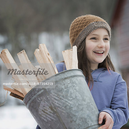 An organic farm in winter in New York State, USA. A girl carrying a bucket full of kindling and firewood.