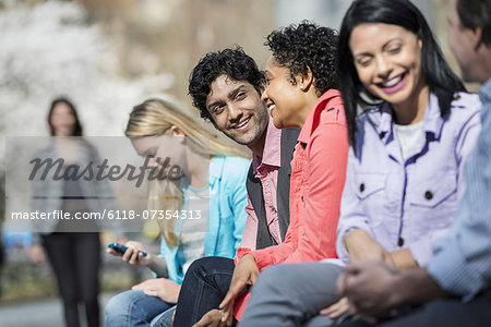 People outdoors in the city in spring time. Five people sitting in a row, one looking at a mobile phone. A woman approaching in the distance. People outdoors in the city in spring time. Five people sitting in a row, one looking at a mobile phone. A woman approaching in the distance.
