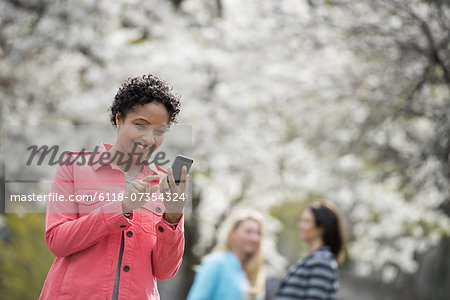 People outdoors in the city in spring time. White blossom on the trees. A young woman checking her cell phone, and laughing.