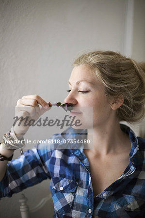 A young woman with blonde hair, holding a aromatic plant or herb flower to her nose and inhaling the aroma.