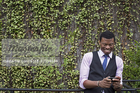 City life in spring. City park with a wall covered in climbing plants and ivy. A young man in a waistcoat, shirt and tie checking his phone. City life in spring. City park with a wall covered in climbing plants and ivy. A young man in a waistcoat, shirt and tie checking his phone.