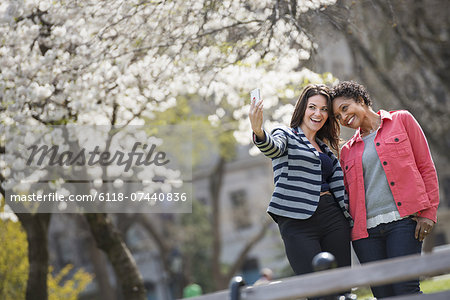 People outdoors in the city in spring time. New York City park. A young woman holding out a phone to take a photograph of herself and a companion.