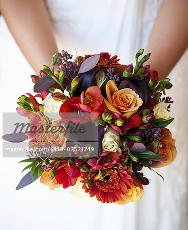 A bride holding a bridal bouquet of colourful red and orange flowers with purple toned leaves.