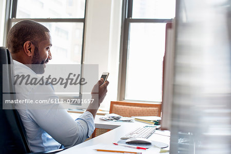 A man working in an office seated at a desk, checking his smart phone.