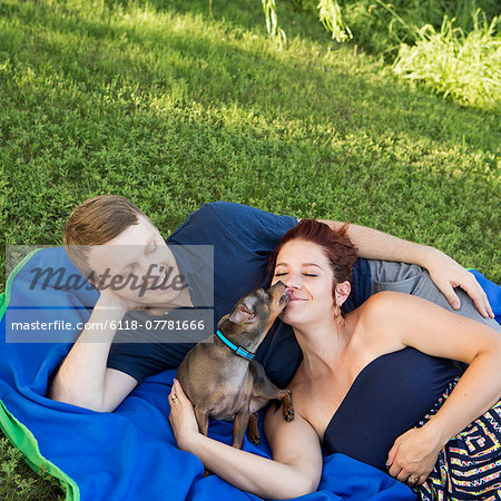 A couple sitting on a picnic rug. A small dog licking the face of a woman.