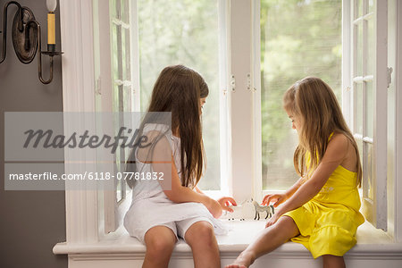 Two girls playing together, sitting on a window seat indoors.