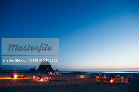 Crowds of people sitting by glowing campfires on Cannon Beach at dusk. Haystack Rock in the background.