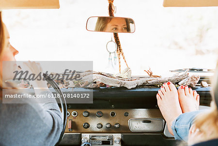 A woman resting her bare feet on the dashboard of a 4x4, on a road trip with another woman driving.