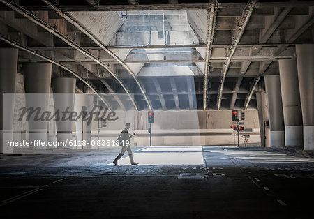A person crossing an open space, a city underpass, with concrete walls and a pool of sunlight.