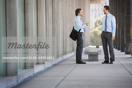 Two businessmen on a walkway outside a building, talking.