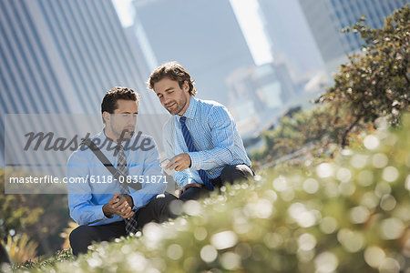 Two men in shirts and ties in a park in the city holding their smart phones.