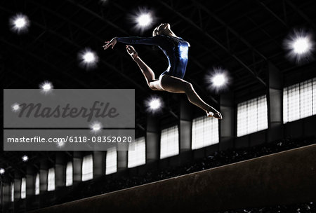 A female gymnast, a young woman performing a floor routine, in mid air with legs and arms outstretched and back bent.