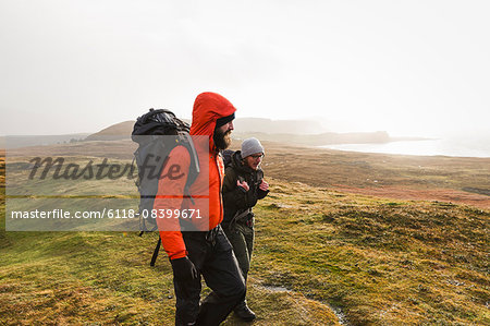 Two men walking with rucksacks, hiking across open ground with a view into the distance of mountains and sea coast.