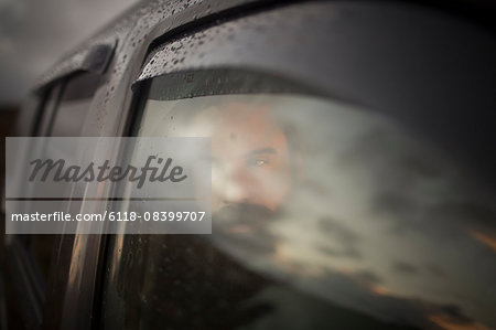 A man sitting in a car looking out. Reflections of the sunset sky on the window.